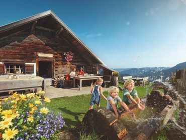 Tre smilende børn leger med vand ved en rustik hytte på en solrig hytteferie med bjerge i baggrunden.