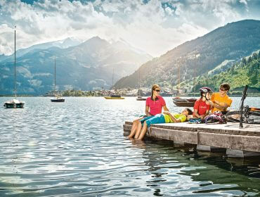 Tre personer i farverigt mountainbikeudstyr slapper af på en kaj ved søen med bjerge og sejlbåde i baggrunden.