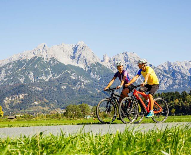 Tauernradweg To personer cykler langs naturskønne cykelruter med grønne marker og bjerge under en klar blå himmel.