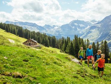 En familie vandrer op ad en græsklædt bjergsti ved en hytte, omgivet af fyrretræer og majestætiske bjerge.