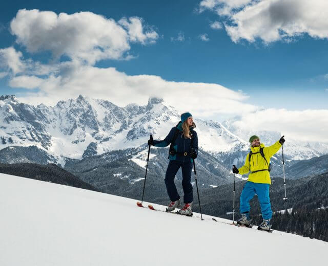 To personer i vintertøj nyder vintervandring, skiløb op ad bakke med sneklædte bjerge og en overskyet himmel bag dem.
