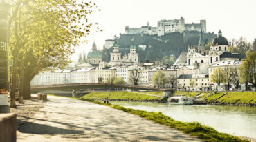 Solrig sti langs floden i Salzburg med fæstningen Hohensalzburg; skisportssteder ligger lige bag de historiske bygninger.