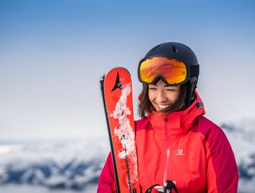 Smilende kvinde i rød skijakke og hjelm holder ski med sneklædte bjerge på et skisportssted i baggrunden.