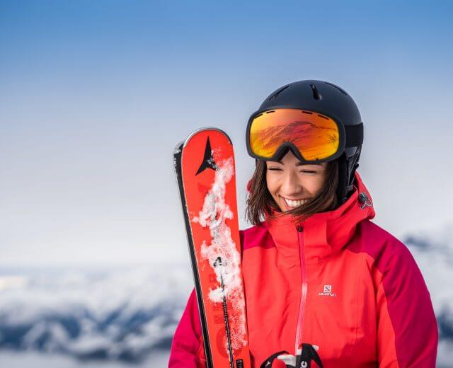 Smilende kvinde i rød skijakke og hjelm holder ski med sneklædte bjerge på et skisportssted i baggrunden.