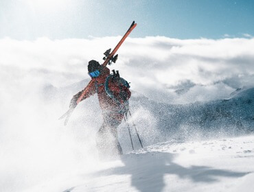 En skiløber i vintertøj bærer ski på skulderen på vej mod den solbeskinnede pist gennem snedækket bjergterræn.