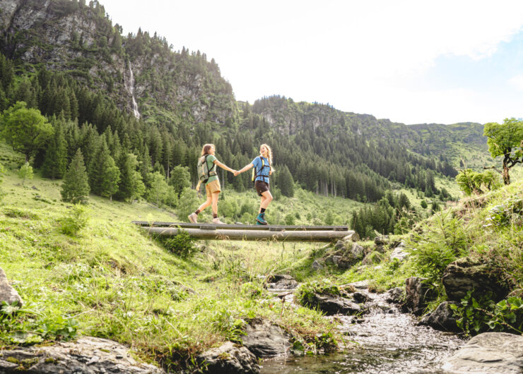 To mennesker holder hinanden i hånden og vandrer over en lille træbro i et frodigt, bjergrigt skilandskab.