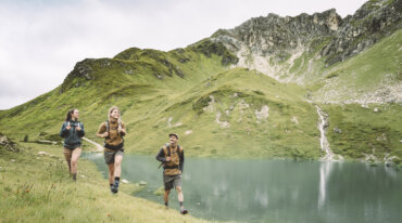 Tre mennesker vandrer langs en græsklædt søbred i nærheden af grønne bjerge, et vandfald og fjerne skiløjper.