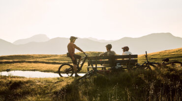 Tre personer med cykler slapper af ved en bænk nær en dam med bjerge, der er værd at stå på ski i baggrunden.