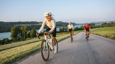 Tre cyklister kører op ad bakke ved en sø på en naturskøn vej i nærheden af marker, træer og fjerne skiløjper.
