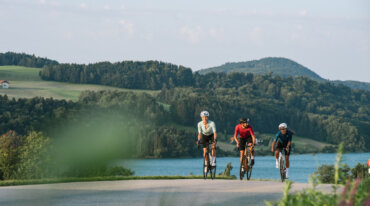 Tre cyklister kører på en landevej nær en sø, hvor folk ofte står på ski om vinteren, under en klar himmel.