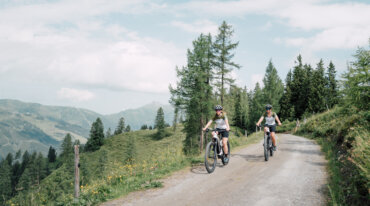 To personer cykler på en grussti gennem et naturskønt, bjergrigt landskab nær skiløjper og vilde blomster.