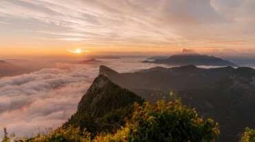 Solopgang over bjerge med skyer i dalene, frodige grønne områder og skiløjper i det fjerne.