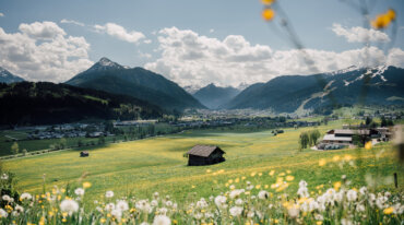 En grøn dal med vilde blomster, en lade, fjerne bjerge og skiløjper under en delvis overskyet himmel.