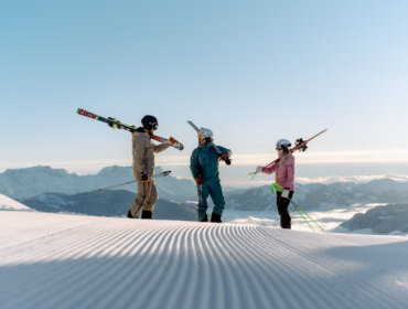 saalbach-winter-skialpin-performer-2024-c-saalbach.com-georg-lindacher-73 Tre skiløbere med udstyr står på en snedækket Skicircus-piste med bjerge og klar himmel i baggrunden.