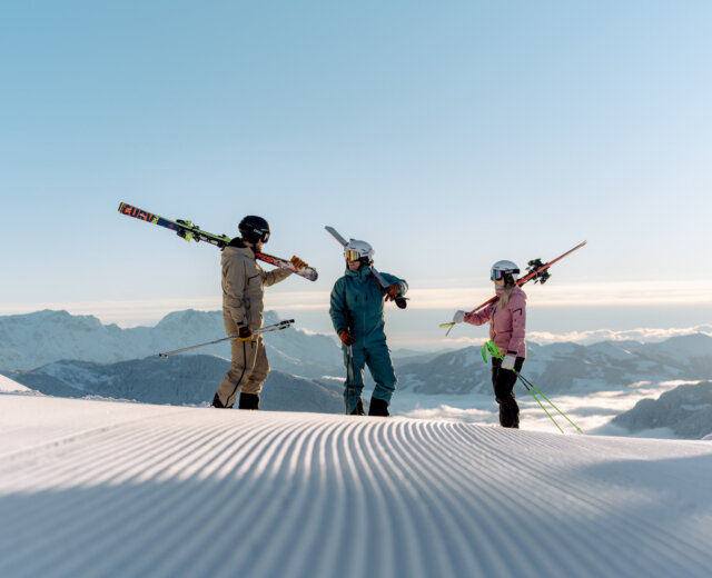 Tre skiløbere med udstyr står på en snedækket Skicircus-piste med bjerge og klar himmel i baggrunden.