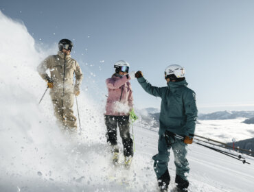 Tre skiløbere i vintertøj giver hinanden knytnæveslag på en snedækket Skicircus-bjergskråning, mens sneen flyver op omkring dem.