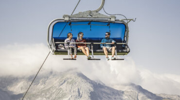Fire personer sidder i en skilift i Obertauern over grønne bakker og bjerge under en blå, skyfyldt himmel.