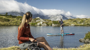 En kvinde sidder ved en sø, mens en mand står på paddleboard; bjerge antyder skiløb i Obertauern bag skyerne.