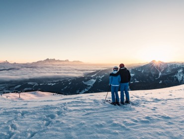 To skiløbere i Wagrain-Kleinarl står på et snedækket bjerg og ser solnedgangen over fjerne tinder og skyer.
