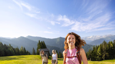 Smilende kvinde på vandretur på en solrig bjergeng med to personer og en østrigsk skihytte i baggrunden.