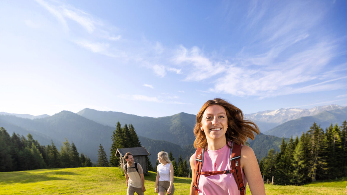 Smilende kvinde vandrer i forgrunden med to personer og et skiklart bjerglandskab under en blå himmel. (Forstørret visning)