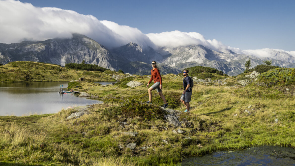 To vandrere står ved en bjergsø og drømmer om at stå på ski i Obertauern under overskyede tinder og en blå himmel. (Forstørret visning)