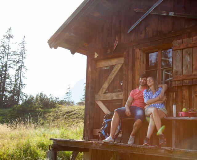 almsommer-huette Et par slapper af på en hytteveranda i den solrige Salzburger Almsommer, omgivet af græsklædte bjerge og træer.