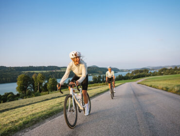 To mennesker cykler på en landevej ved en sø, omgivet af grønne bakker under en klar blå himmel.