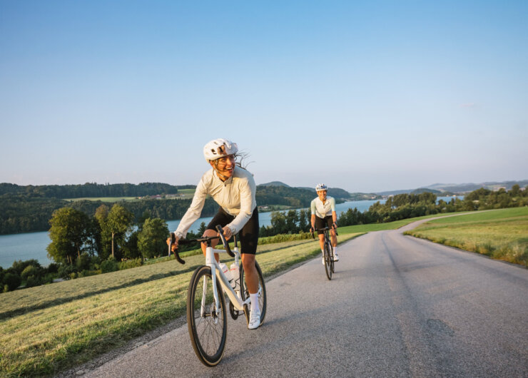 To mennesker cykler på en landevej ved en sø, omgivet af grønne bakker under en klar blå himmel.