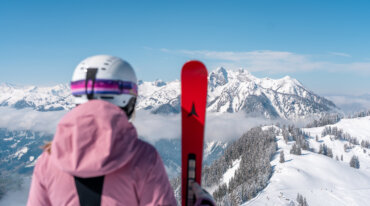 Person i lyserød jakke og hjelm med rød ski i hånden med udsigt til det sneklædte St. Johann i Salzburgs bjerge.