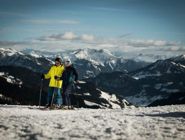 To personer i skitøj står på en sneklædt skråning med snedækkede bjerge i baggrunden.
