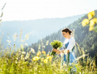 Kvinde på en mark med en buket vilde blomster og bjerge i baggrunden på en solrig dag.