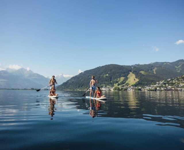 c-zell-am-see-kaprun-tourismus To personer på paddleboard på en rolig sø med bjerge og en by i baggrunden under en klar himmel.