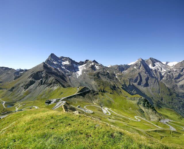 Snoet bjergvej med en rød bil, grønne bakker og sneklædte tinder under en klar blå himmel.