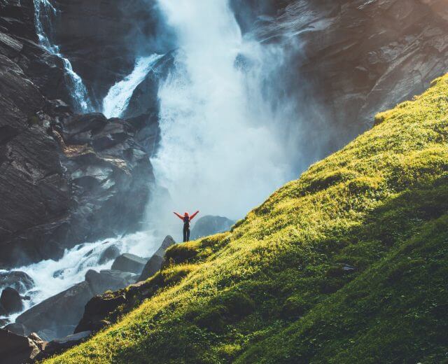 En person med løftede arme står på en klippe nær et højt vandfald og en frodig, grøn bjergside i sollys.