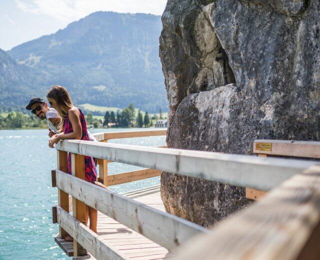 To mennesker står på en træbro over en sø med bjerge og træer i baggrunden.