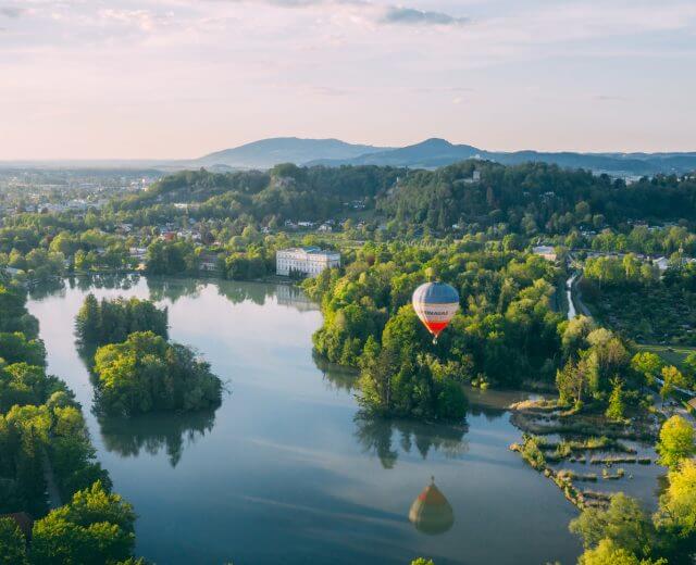 En varmluftballon svæver over en sø omgivet af træer og bakker under en delvis overskyet himmel.