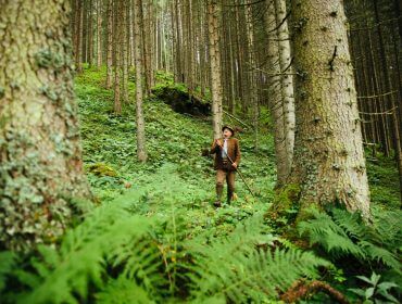 En person i brunt tøj og hat står med en spadserestok i en frodig, grøn skov omgivet af høje træer.