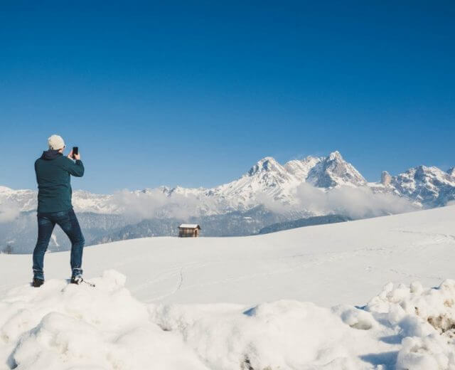 En person i vintertøj tager et billede af snedækkede bjerge under en klar blå himmel.