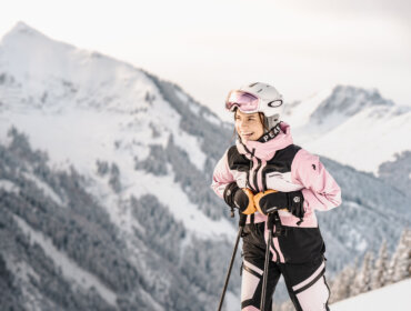 Smilende skiløber i pink og sort udstyr står på et snedækket bjerg med skistave; snedækkede tinder i baggrunden.