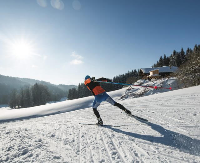 Person på langrendsski på en snedækket løjpe med træer og en hytte under en lys, solrig himmel.