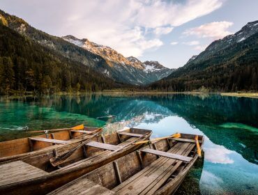 4-Wagrain Robåde af træ på en klar bjergsø med fyrretræer og snedækkede tinder i baggrunden.