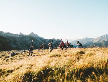 En gruppe mennesker laver yogastillinger på en græsklædt bakke med bjerge i baggrunden.