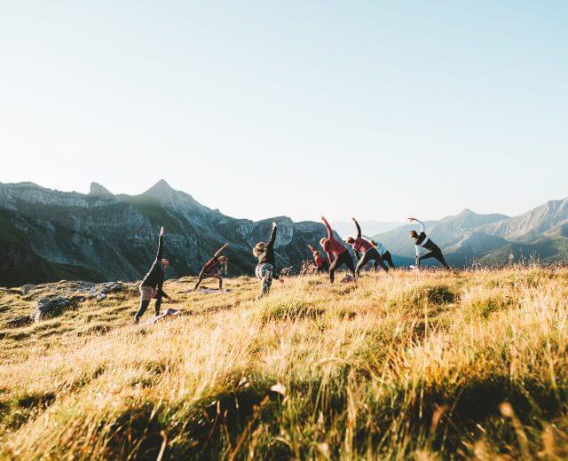 _DSC0256 En gruppe mennesker laver yogastillinger på en græsklædt bakke med bjerge i baggrunden.