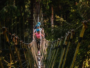 Tre personer iført hjelme krydser en rebbro i en eventyrpark i skoven.