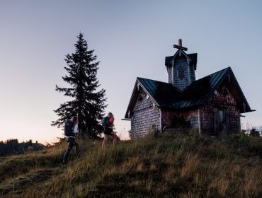To vandrere nærmer sig en lille trækirke i skumringen i SalzburgerLand med et højt fyrretræ i nærheden.