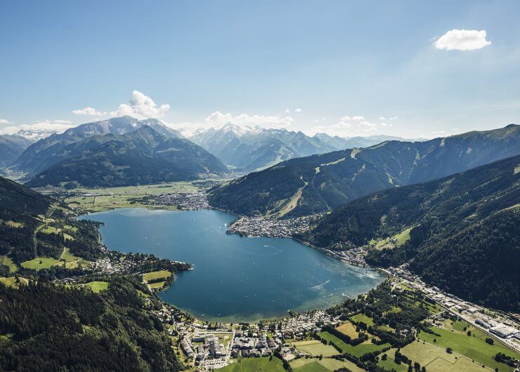 Luftfoto af en sø omgivet af grønne bakker, bjerge og en lille by under en klar blå himmel.