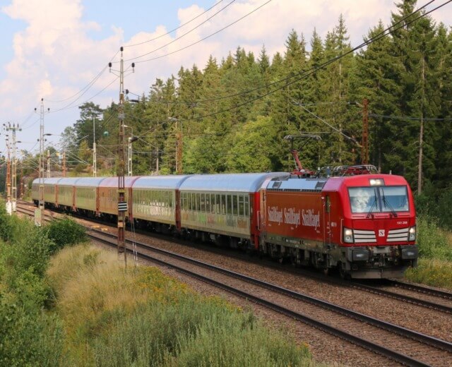 Et rødt passagertog kører på skinner gennem et grønt, skovklædt område under en delvis overskyet himmel.