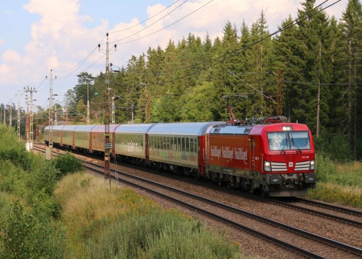 Et rødt passagertog kører på skinner gennem et grønt, skovklædt område under en delvis overskyet himmel.