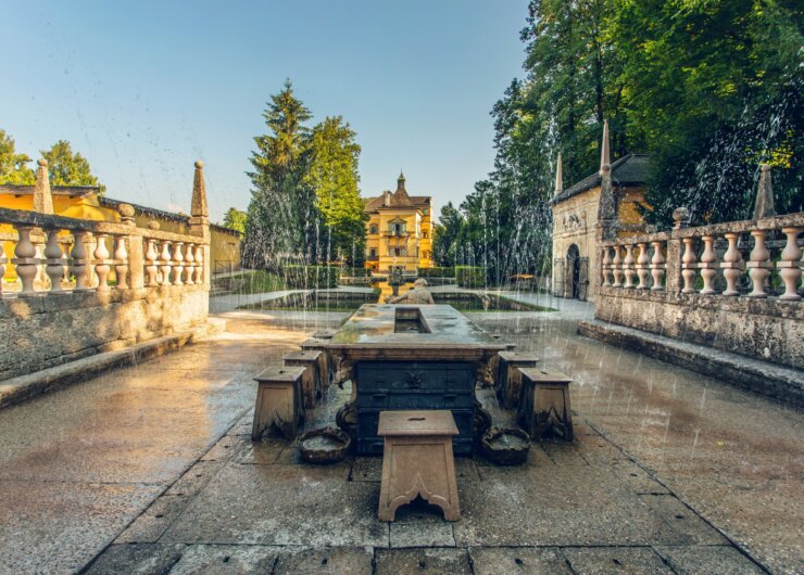 Stensti med springvand og en gul villa i SalzburgerLand, omgivet af træer under en klar blå himmel.